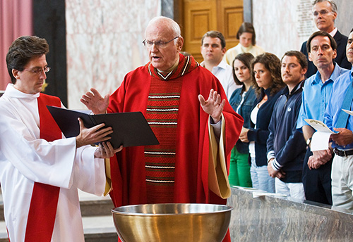 Priest in red blessing a basin of water with a deacon holding the ritual book