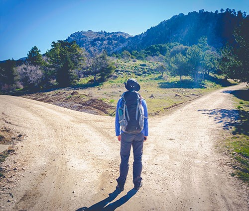 Person with a backpack standing at a split in a dirt road as if choosing a route